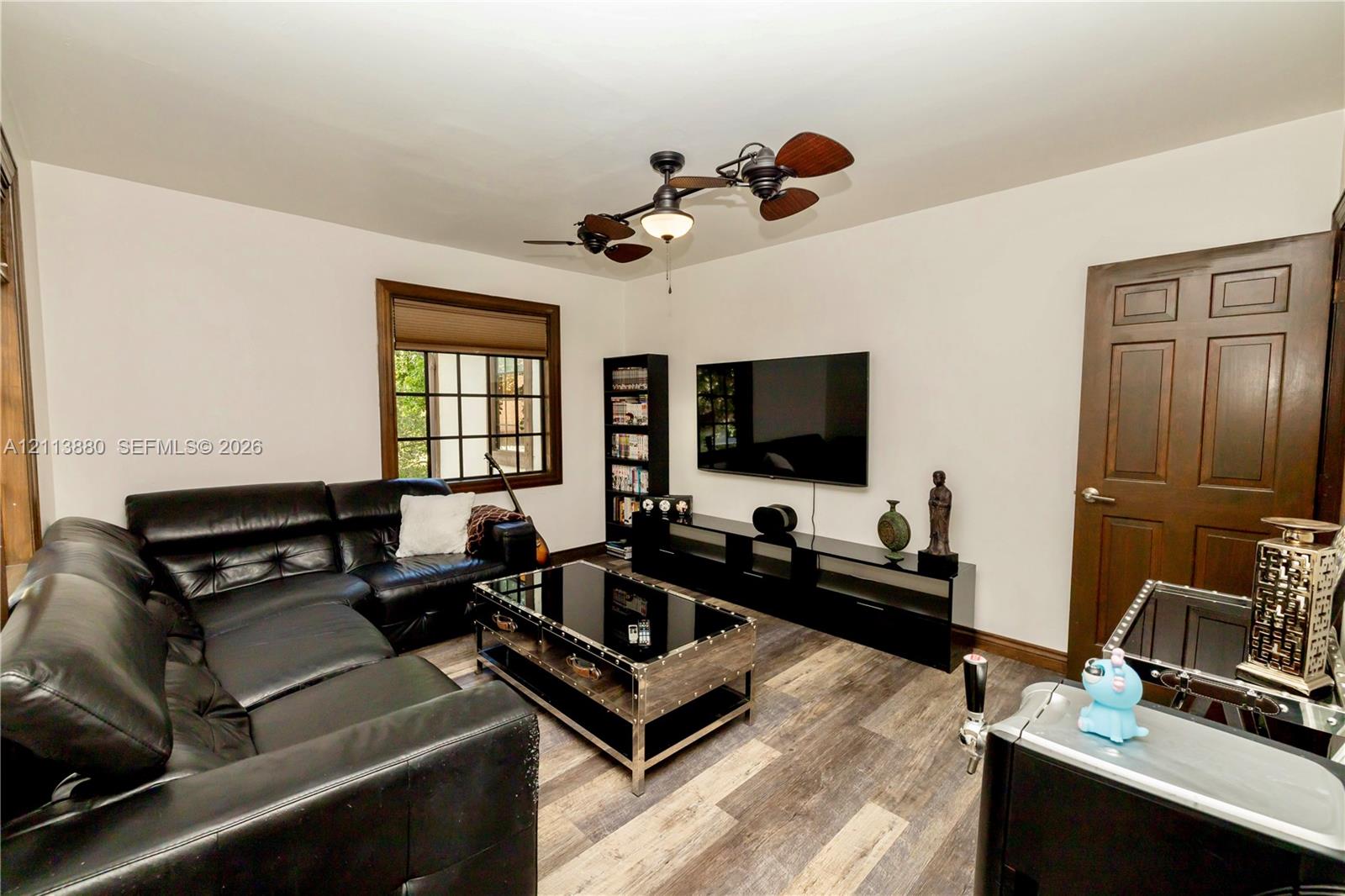 Multi-tonal wood-finish flooring, dark-stained six-panel door, multi-pane window with dark-stained trim, and multi-blade ceiling fan with integrated lighting