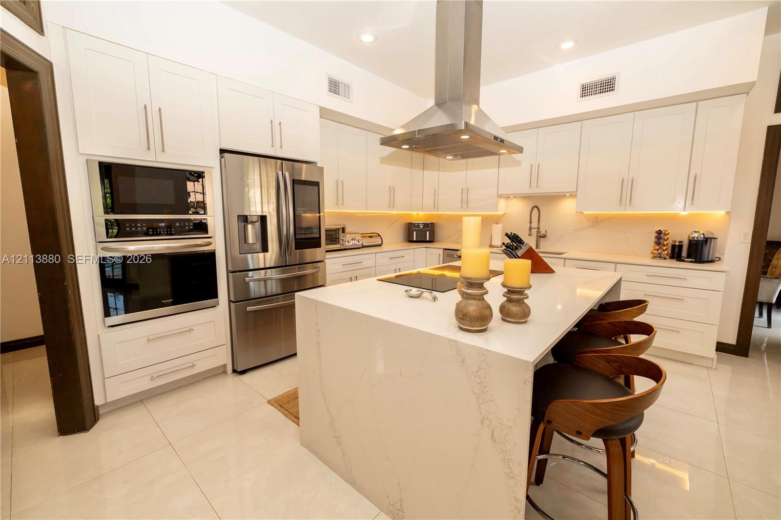 Kitchen featuring white cabinetry, quartz countertops with a waterfall edge island, stainless steel wall-mounted range hood, built-in stainless steel double oven, and integrated cabinet lighting