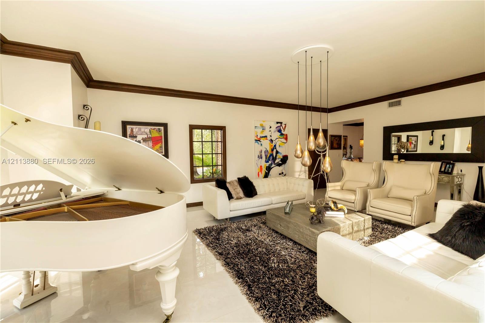 Spacious living area featuring polished tile flooring, prominent crown molding, multi-pendant tiered lighting, and large-paned windows with dark wood trim