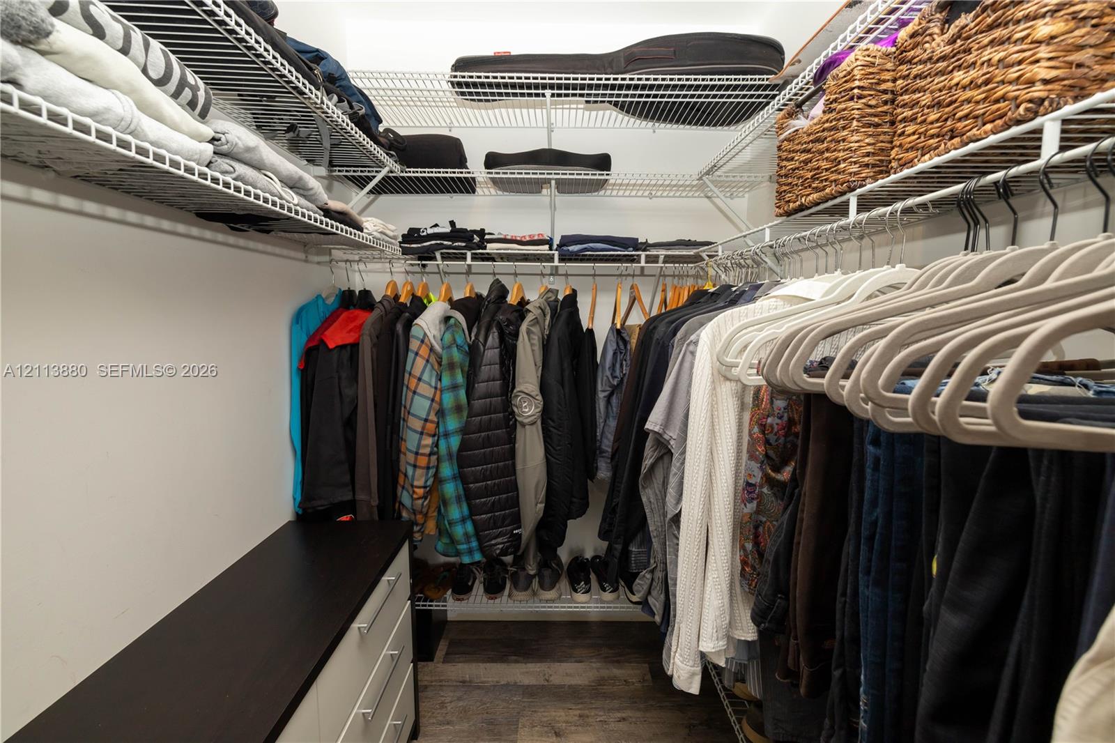 Walk-in closet featuring multi-tiered wire shelving, integrated hanging rods, and a built-in cabinet with a dark-finish countertop over wood-finish flooring