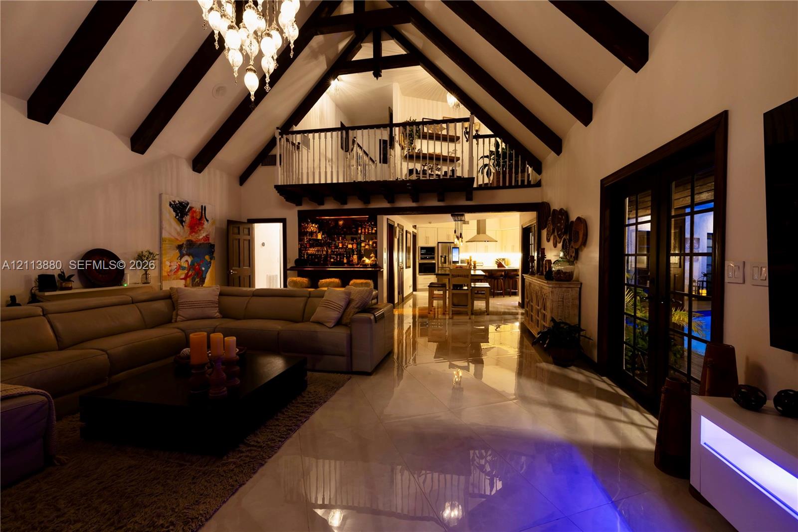 Vaulted ceilings with exposed timber beams, polished stone flooring, custom ornamental iron chandelier, built-in wet bar, and lofted mezzanine with spindle railing