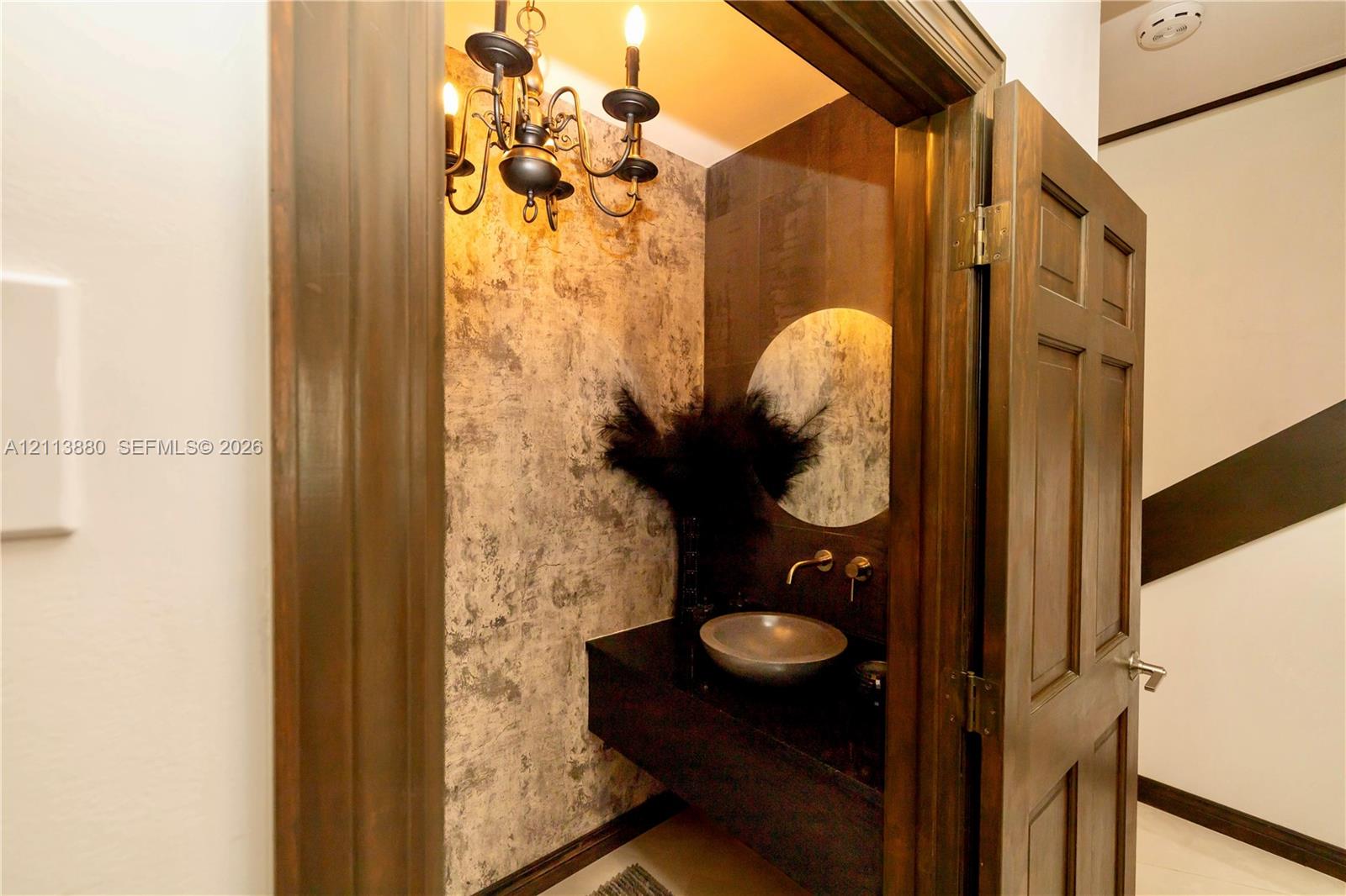 Textured wall covering, floating vanity with a basin-style sink, wall-mounted faucet, round mirror, and traditional chandelier