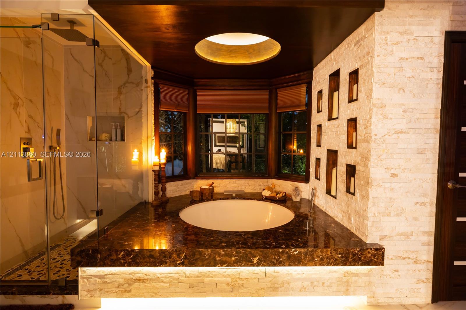 Circular soaking tub set into a stone-finish deck with under-cabinet lighting, glass-enclosed shower with large-format wall tiling, recessed wall shelving, and bay window arrangement