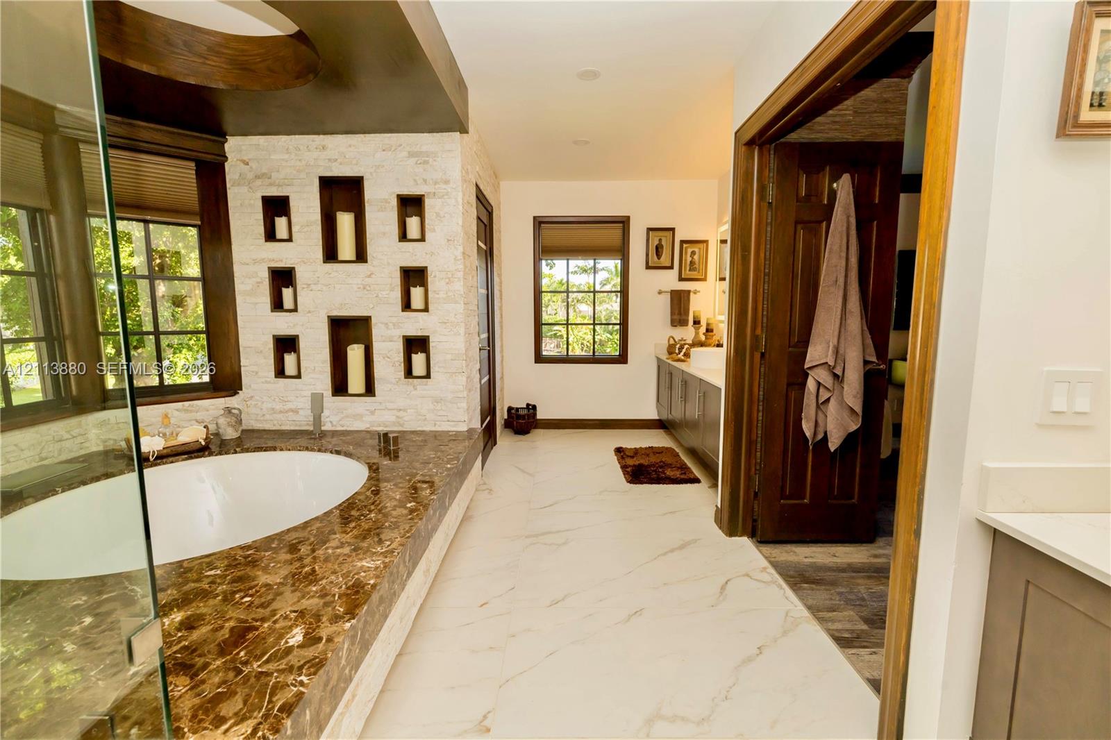 Oversized soaking tub with stone surround, textured stone feature wall with integrated display niches, large-format tile flooring, and dark wood-finished cabinetry
