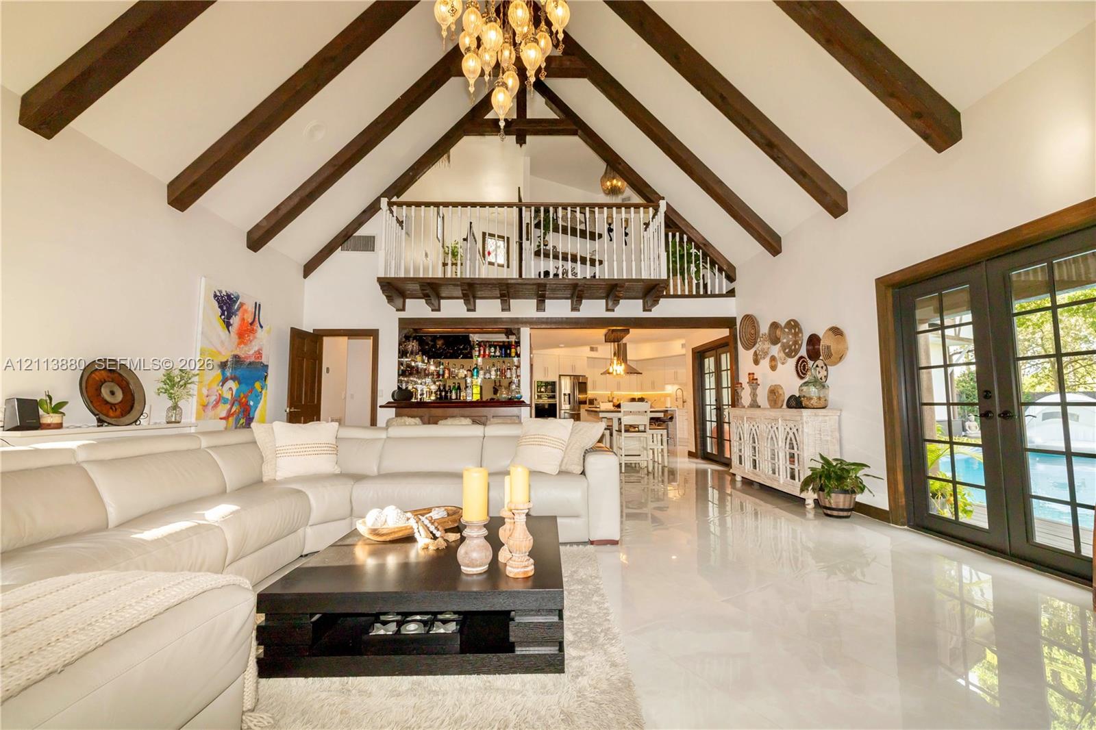 Vaulted ceiling with exposed wooden beams, interior wooden railed loft, polished stone flooring, multi-light hanging chandelier, and dual-pane glass French doors