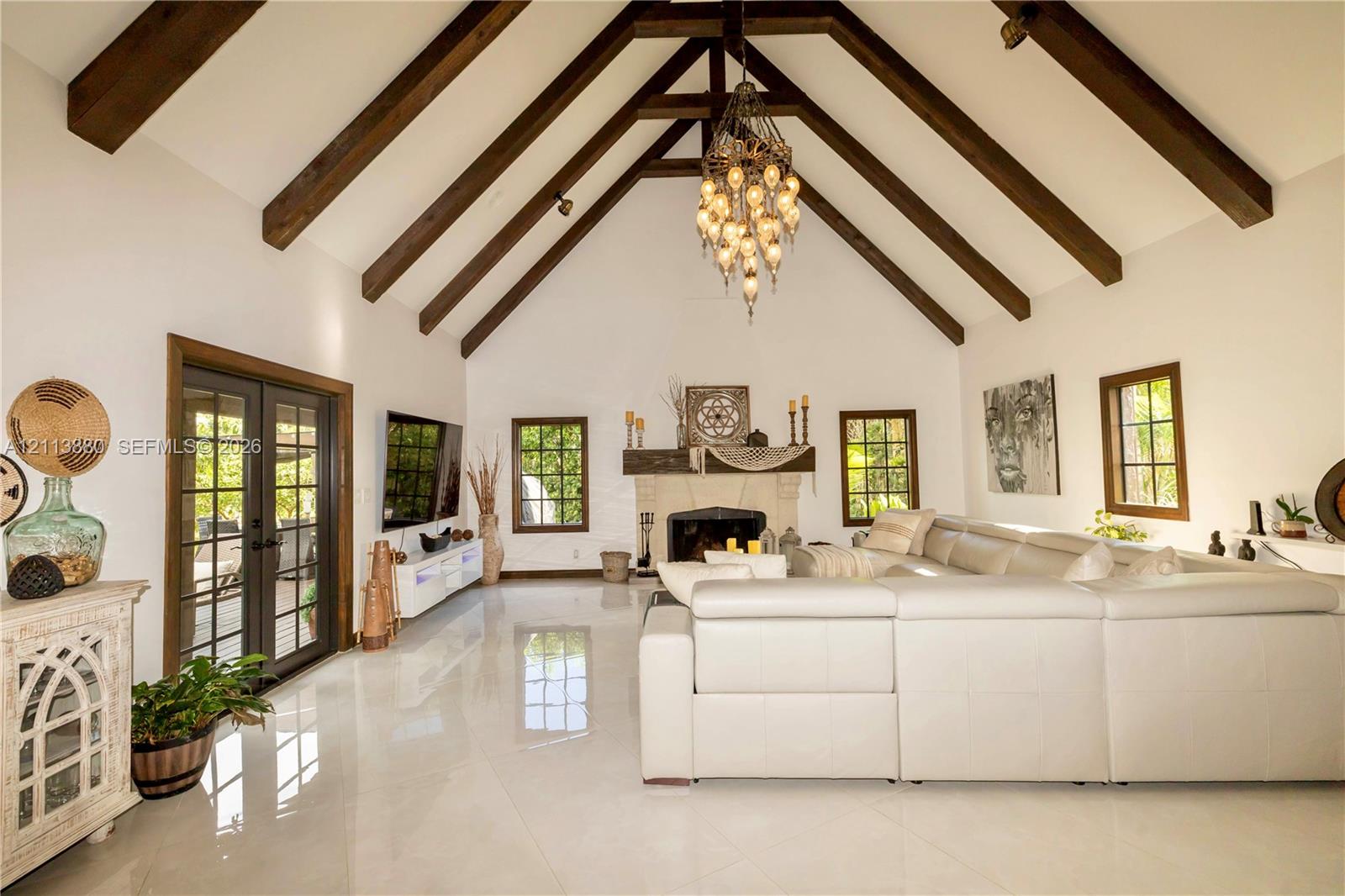 Vaulted ceiling with exposed wood-finish beams, polished tile flooring, stone-surround fireplace with a wooden mantel, and multi-pane glass french doors