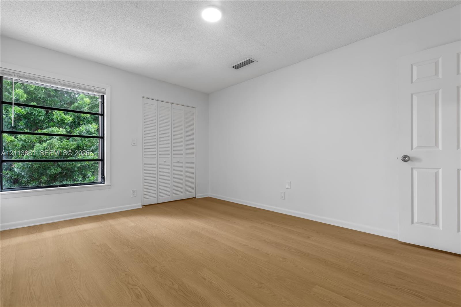 Wood-finish flooring, large window with natural scenery, ceiling-mounted light fixture, and white louvered closet doors