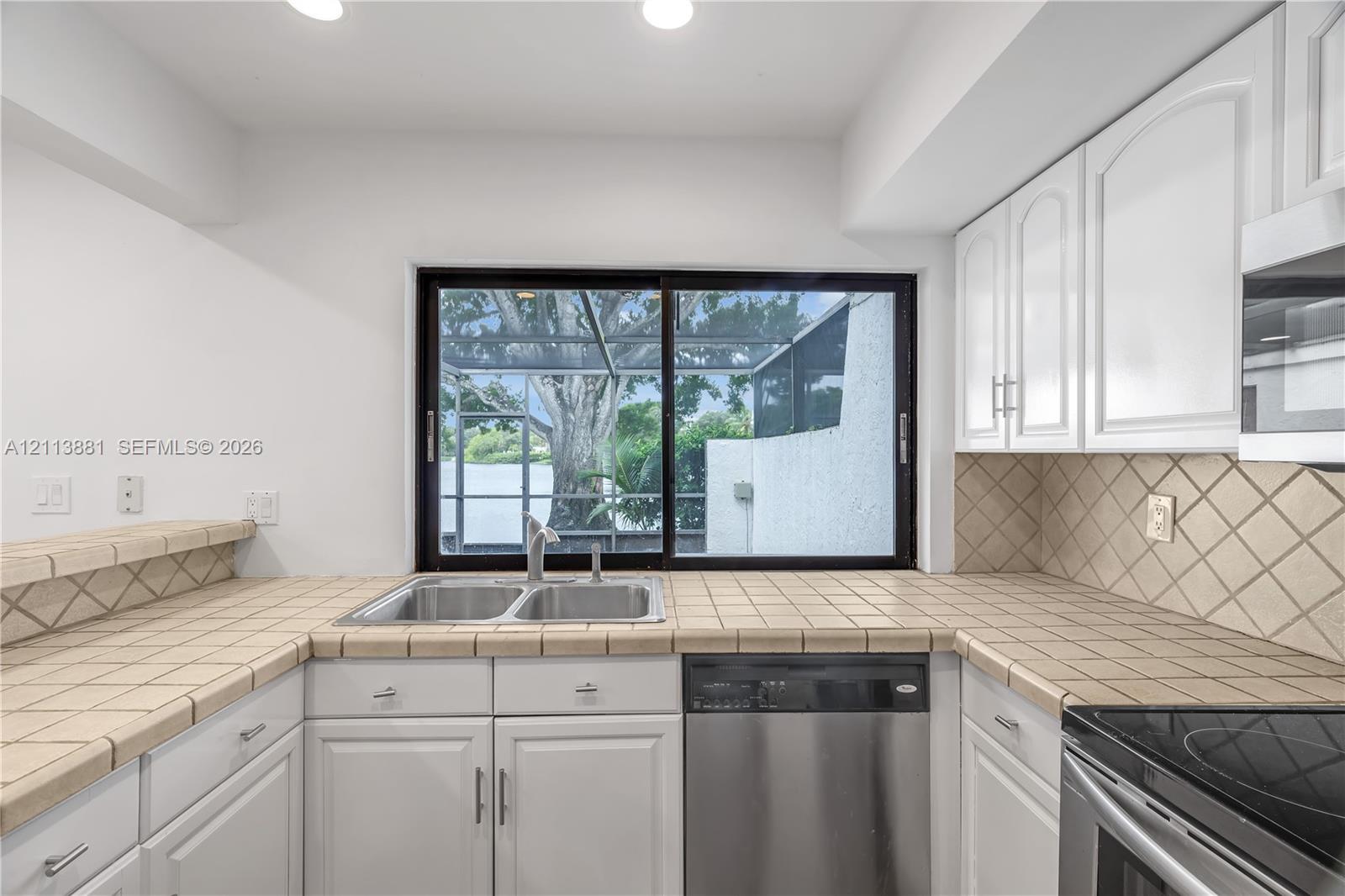 Kitchen featuring white cabinetry, tile countertops, diamond-patterned tile backsplash, stainless steel dishwasher, and sliding glass window