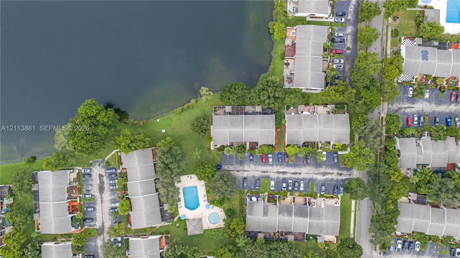 Aerial perspective of multi-family residences featuring waterfront positioning, expansive green space, mature trees, and a shared swimming pool
