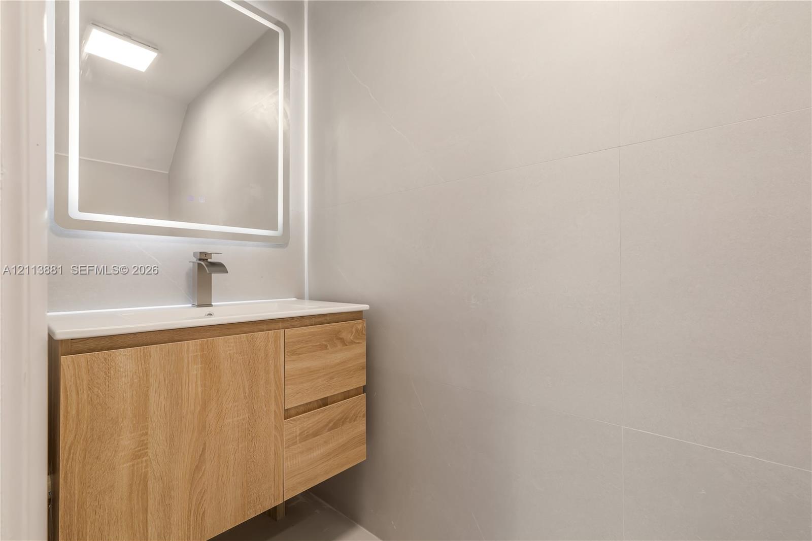 Floating wood-finish vanity with a rectangular sink, brushed nickel faucet, wall-mounted LED-backlit mirror, and large-format neutral wall tile
