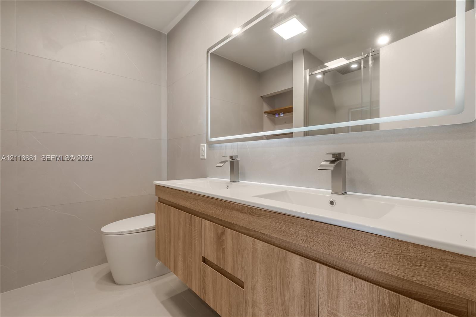 Floating wood-finish vanity with dual integrated sinks, wall-mounted metallic faucets, large backlit mirror, and floor-to-ceiling tiled walls