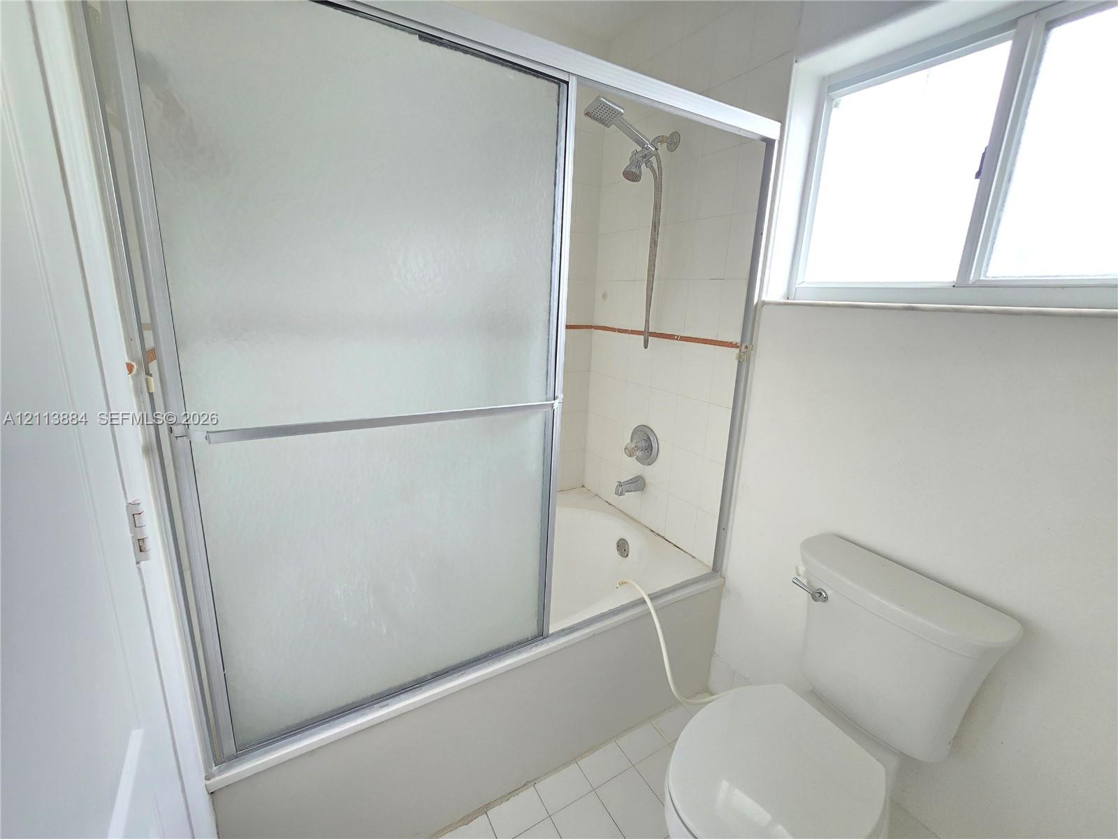 Bathtub and shower combination featuring sliding textured glass doors, tiled surround, wall-mounted shower head, and white floor tiling