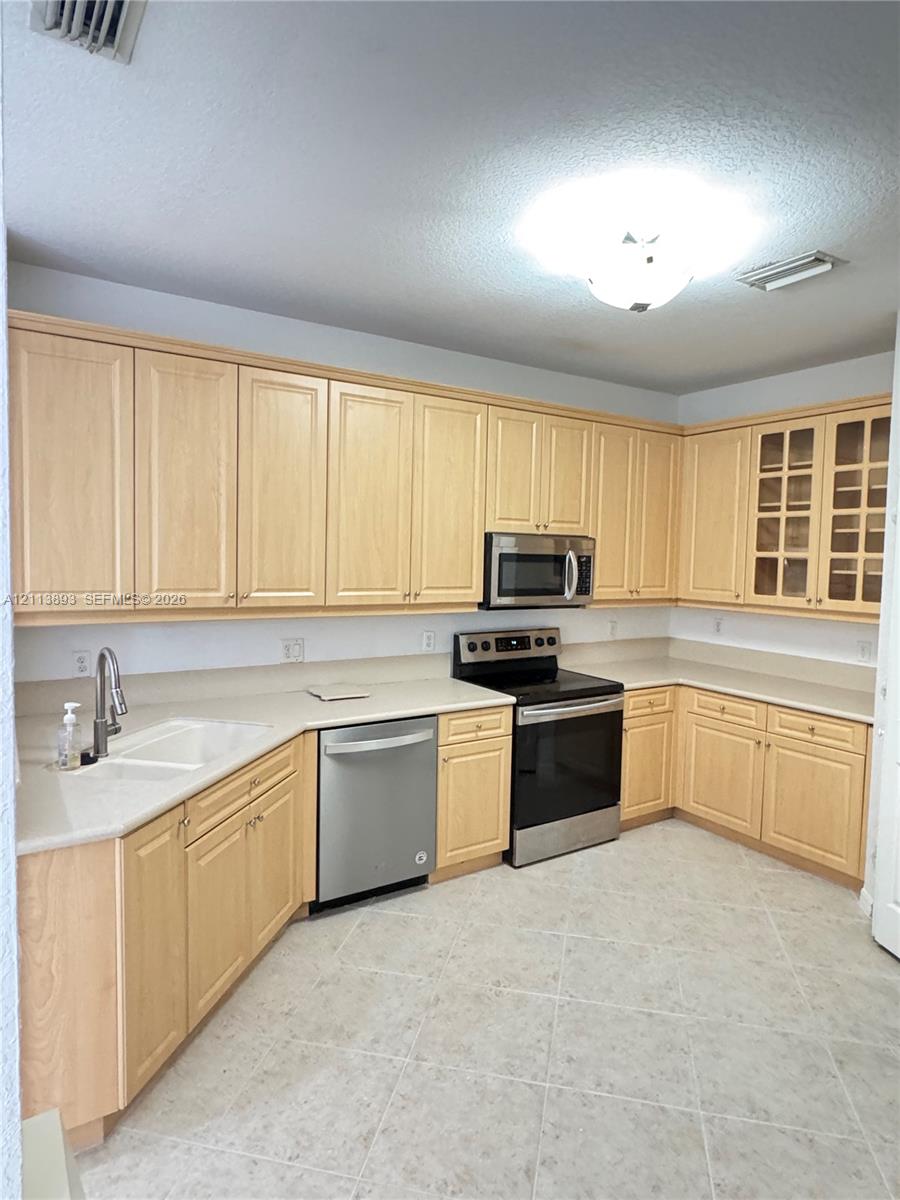 Light-toned cabinetry with integrated glass-paneled display uppers, expansive solid-surface countertops, tile flooring, and stainless steel appliances including a built-in microwave