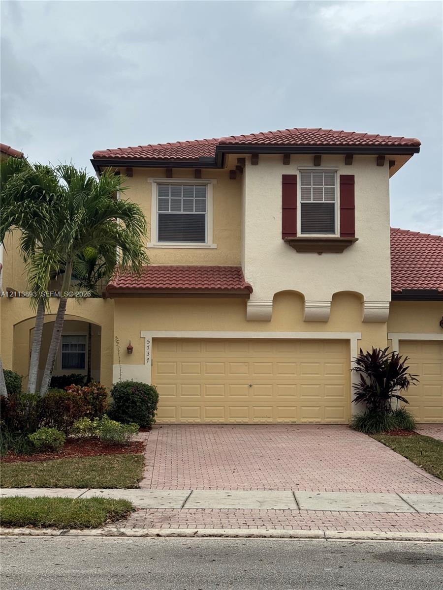 Stucco exterior featuring a terracotta tile roof, decorative red shutters, arched corbels, and a paved driveway