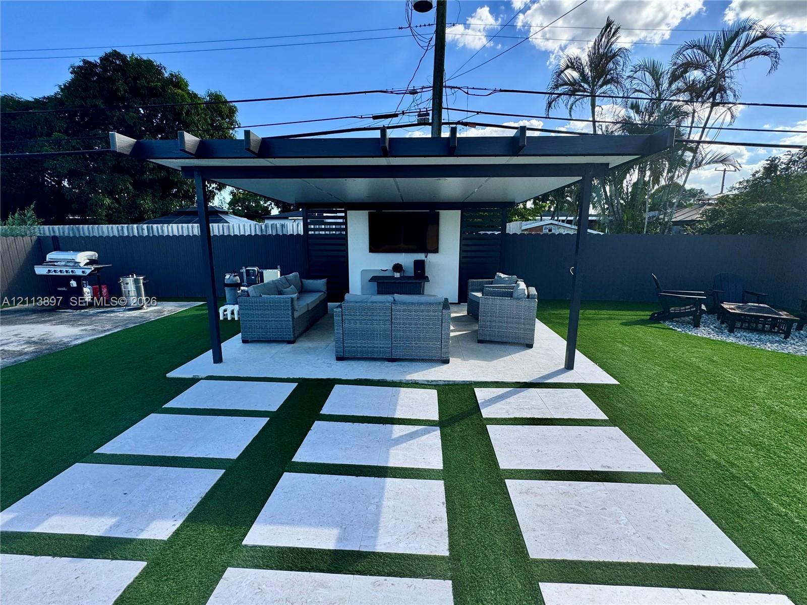 Outdoor covered pavilion with a solid roof structure, concrete paver pathway, synthetic turf lawn, and perimeter privacy fencing