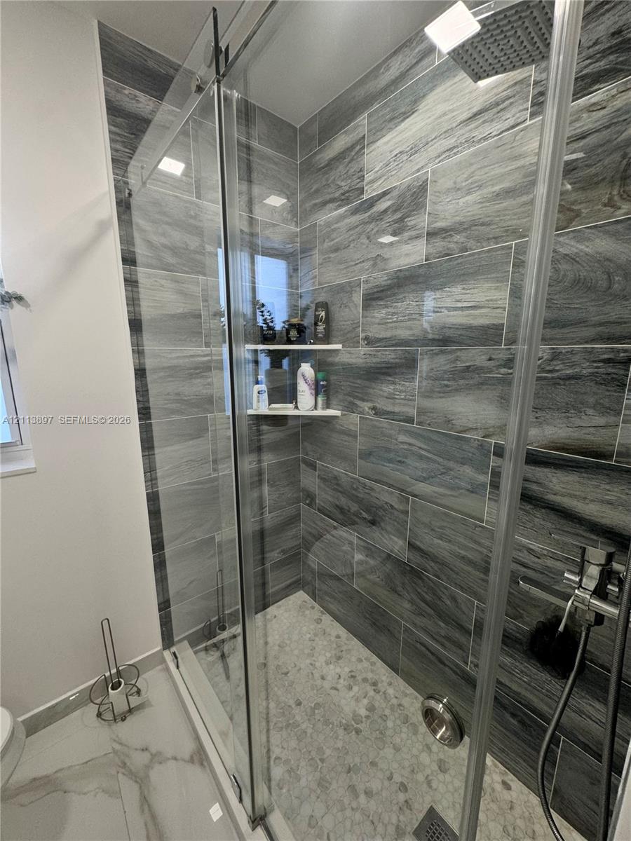 Walk-in shower featuring large-format stone-pattern wall tile, pebble-texture floor tiling, a frameless glass sliding door, a built-in corner shelf, and a rainfall showerhead