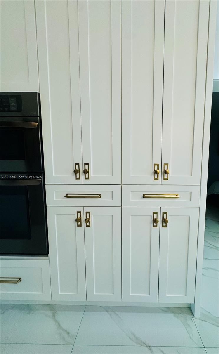 White shaker-style cabinetry with recessed panels, brushed brass hardware, and a built-in double wall oven, set against large-format rectified tile flooring