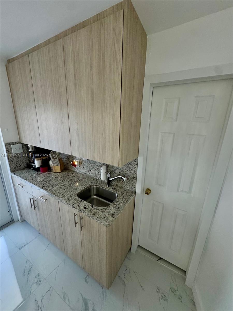 Flat-panel wood-finish cabinetry, speckled stone countertop, stainless steel sink with high-arc faucet, and large-format tile flooring