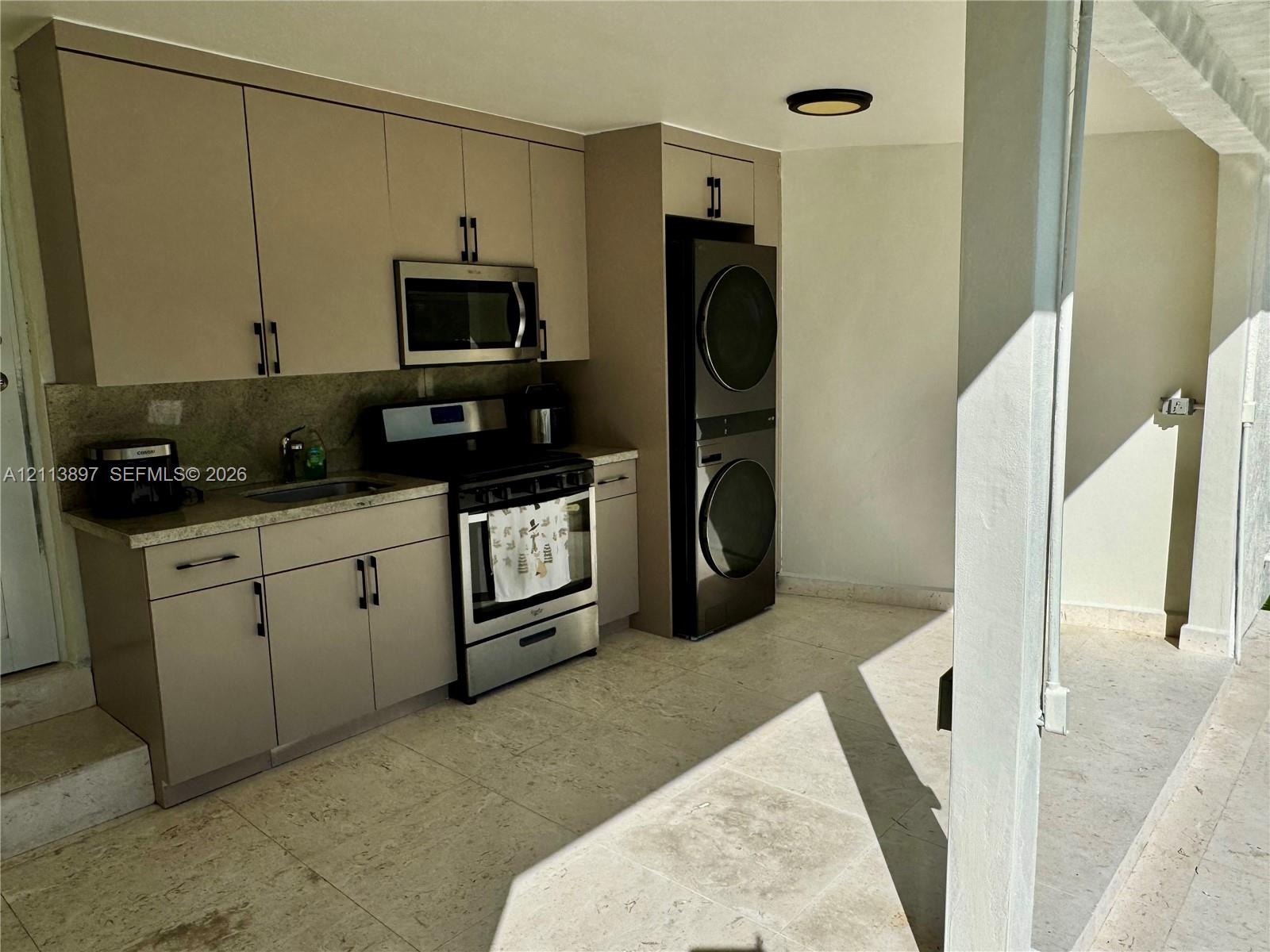 Kitchenette featuring flat-panel cabinetry, stone-finish countertops, stainless steel appliances, and stacked laundry unit, complemented by tile flooring