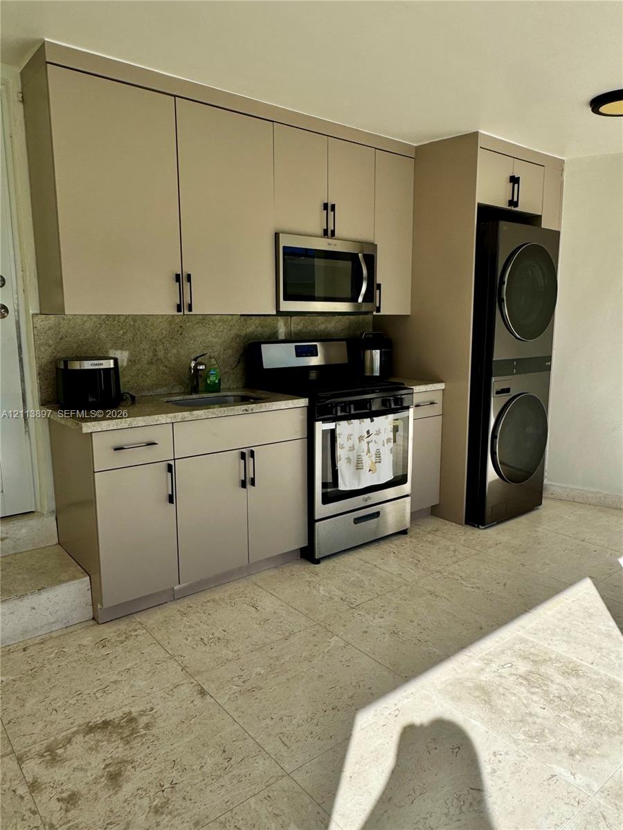 Kitchenette featuring flat-panel cabinetry, stone countertops, stainless steel appliances, and a stacked laundry unit set on stone-finish flooring