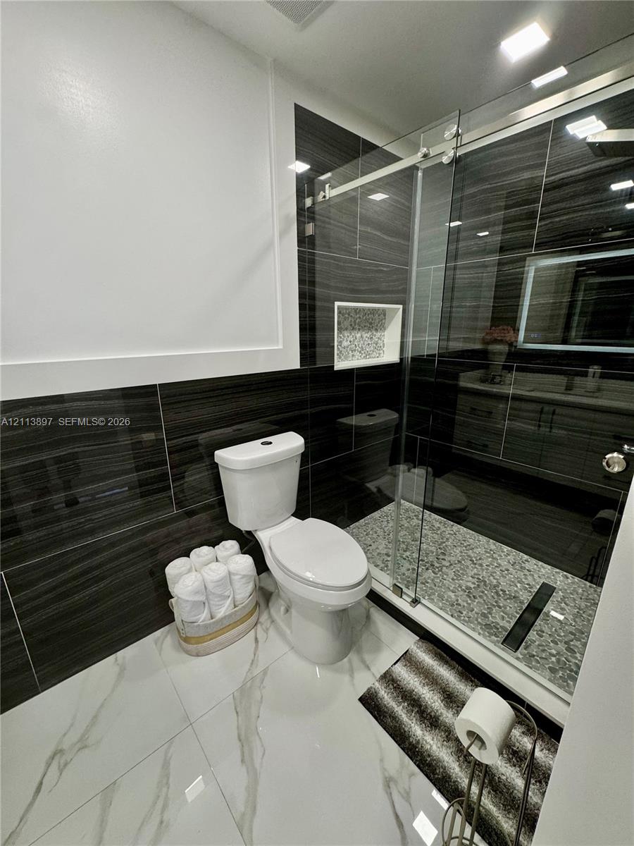 Bathroom featuring large-format white tile flooring, dark horizontal-patterned wall tiling, a walk-in shower with a glass sliding door, a recessed shower niche, and a vanity cabinet with an integrated LED-lit mirror