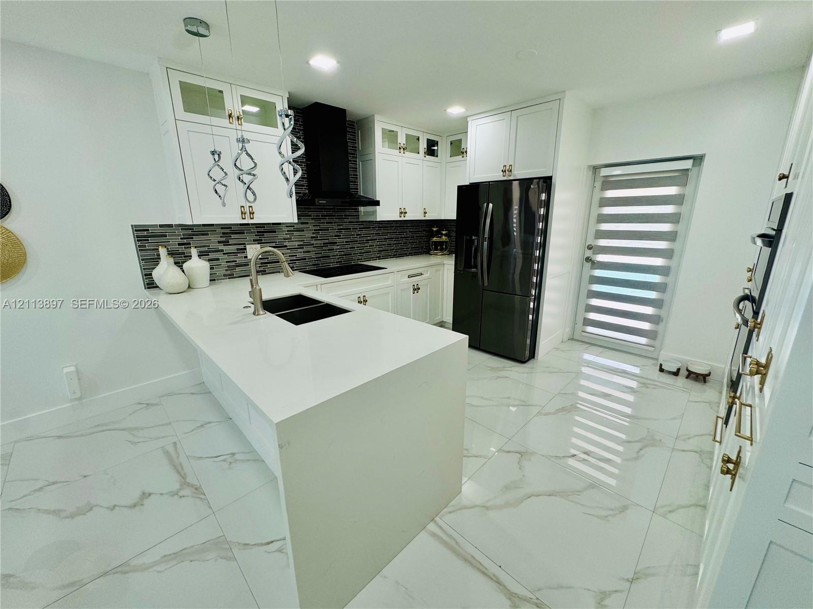 Kitchen featuring white cabinetry with glass-panel accents, stone-composite countertops, a multi-tonal linear tile backsplash, polished stone-finish flooring, and a central island with a double-basin under-mount sink