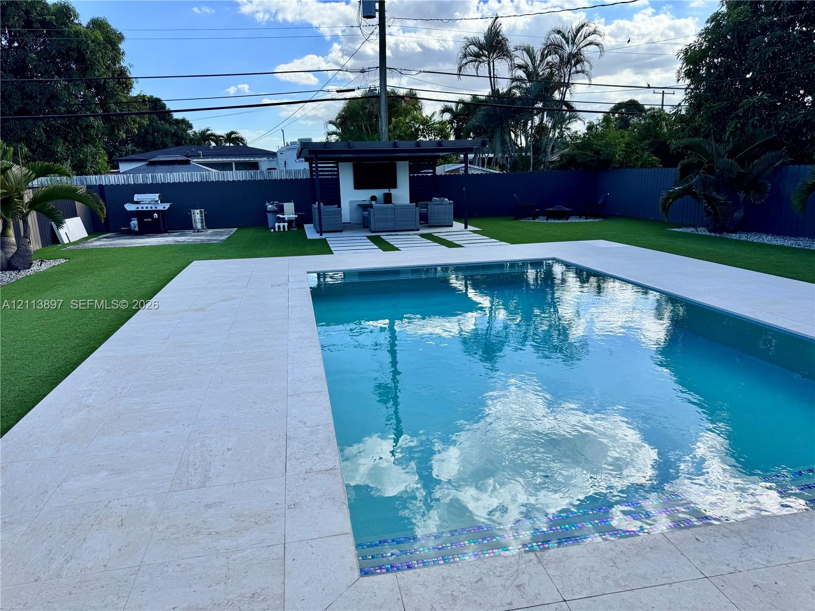 In-ground pool with stone tile surround, covered patio structure, synthetic lawn, and perimeter privacy fencing