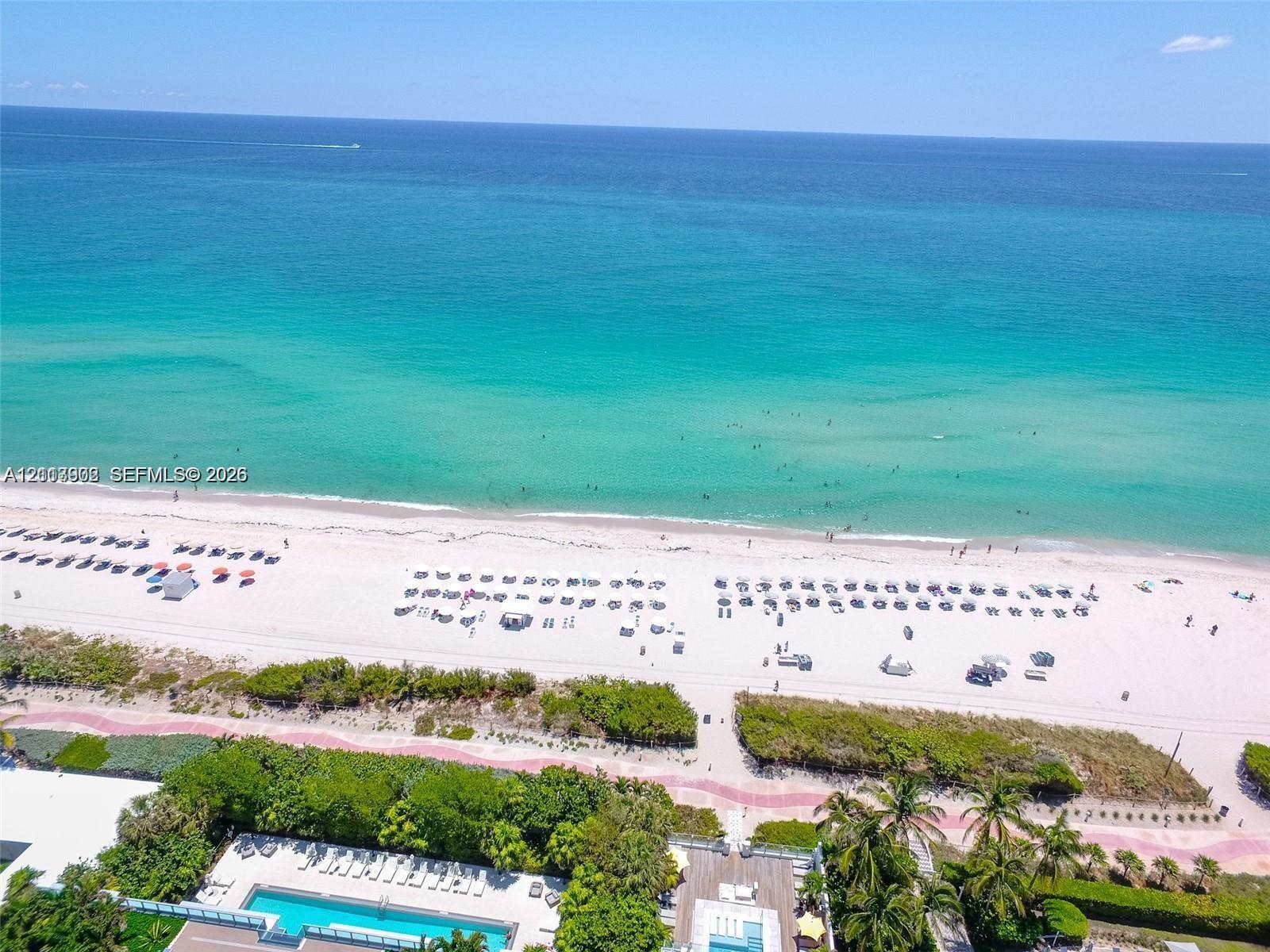 Direct oceanfront access with expansive white sand beach, paved coastal pedestrian pathway, and landscaped grounds featuring a rectangular swimming pool with integrated sun deck