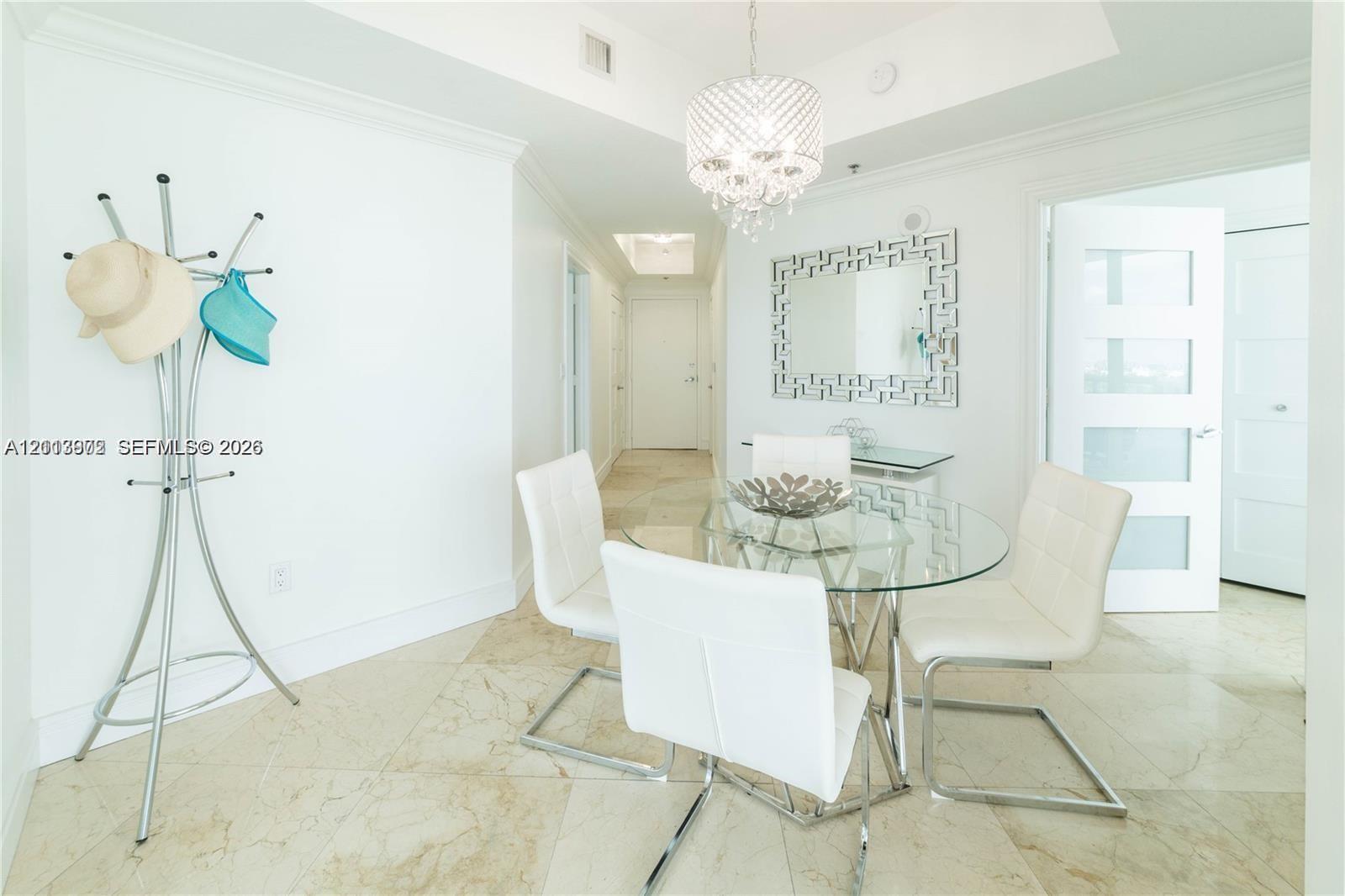 Light-toned stone tile flooring throughout, recessed tray ceiling, crystal-style chandelier, and white-framed door with translucent panels