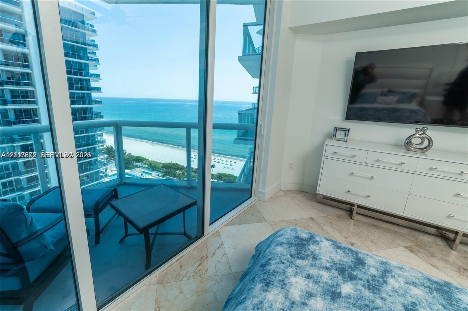 Floor-to-ceiling glass walls, private balcony with glass railing, stone flooring, and expansive ocean and shoreline scenery