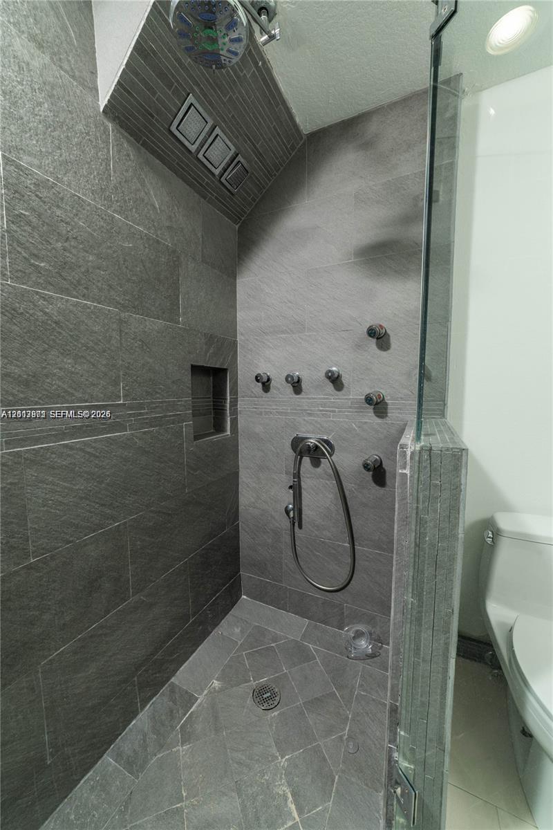 Walk-in shower featuring large-format dark grey stone tile walls, a built-in recessed niche, multiple body sprayers, a handheld shower head, and a glass partition