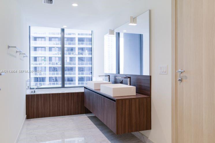 Floating wood-finish double vanity with dual square vessel sinks and chrome faucets, large frameless wall mirror, soaking tub with matching wood-finish surround, floor-to-ceiling windows, and light-toned stone-finish flooring