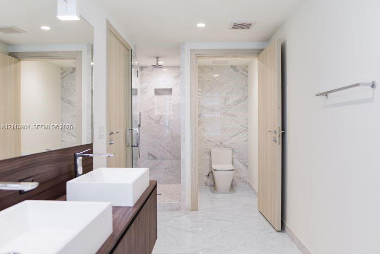 Dual vessel sinks atop a wood-finish vanity, stone-surround walk-in shower with integrated niche, stone flooring, and polished chrome fixtures