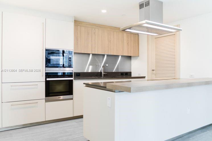 Modern kitchen featuring white flat-panel cabinetry, wood-finish upper cabinetry, a stainless steel range hood, integrated oven and microwave, and a stone-surface island with a breakfast bar