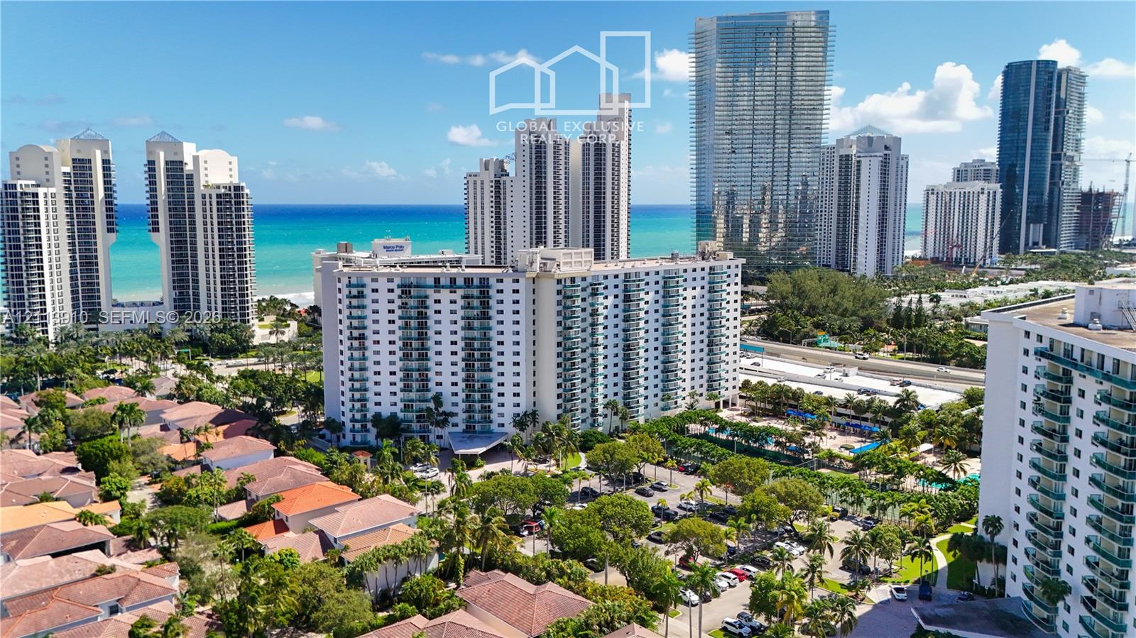 Aerial perspective featuring a multi-story residential building with private balconies, surrounded by mature palm trees, paved parking areas, and proximity to the coastline Aerial perspective featuring a multi-story residential building with private balconies, surrounded by mature palm trees, paved parking areas, and proximity to the coastline