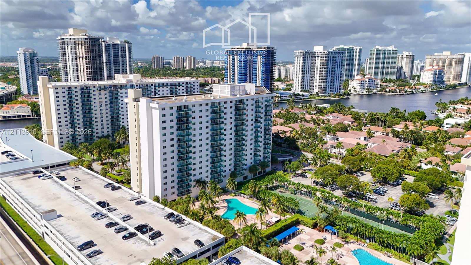 Aerial perspective of a multi-story residential complex featuring an outdoor swimming pool, tennis courts, paved parking structures, and mature landscaping Aerial perspective of a multi-story residential complex featuring an outdoor swimming pool, tennis courts, paved parking structures, and mature landscaping