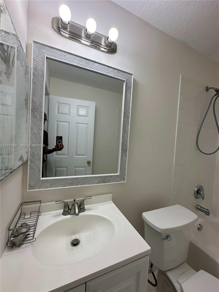 Bathroom featuring a vanity with a white countertop and oval sink, a wall-mounted mirror with a textured frame, a three-light vanity fixture, and a tub-shower combination with tiled surround
