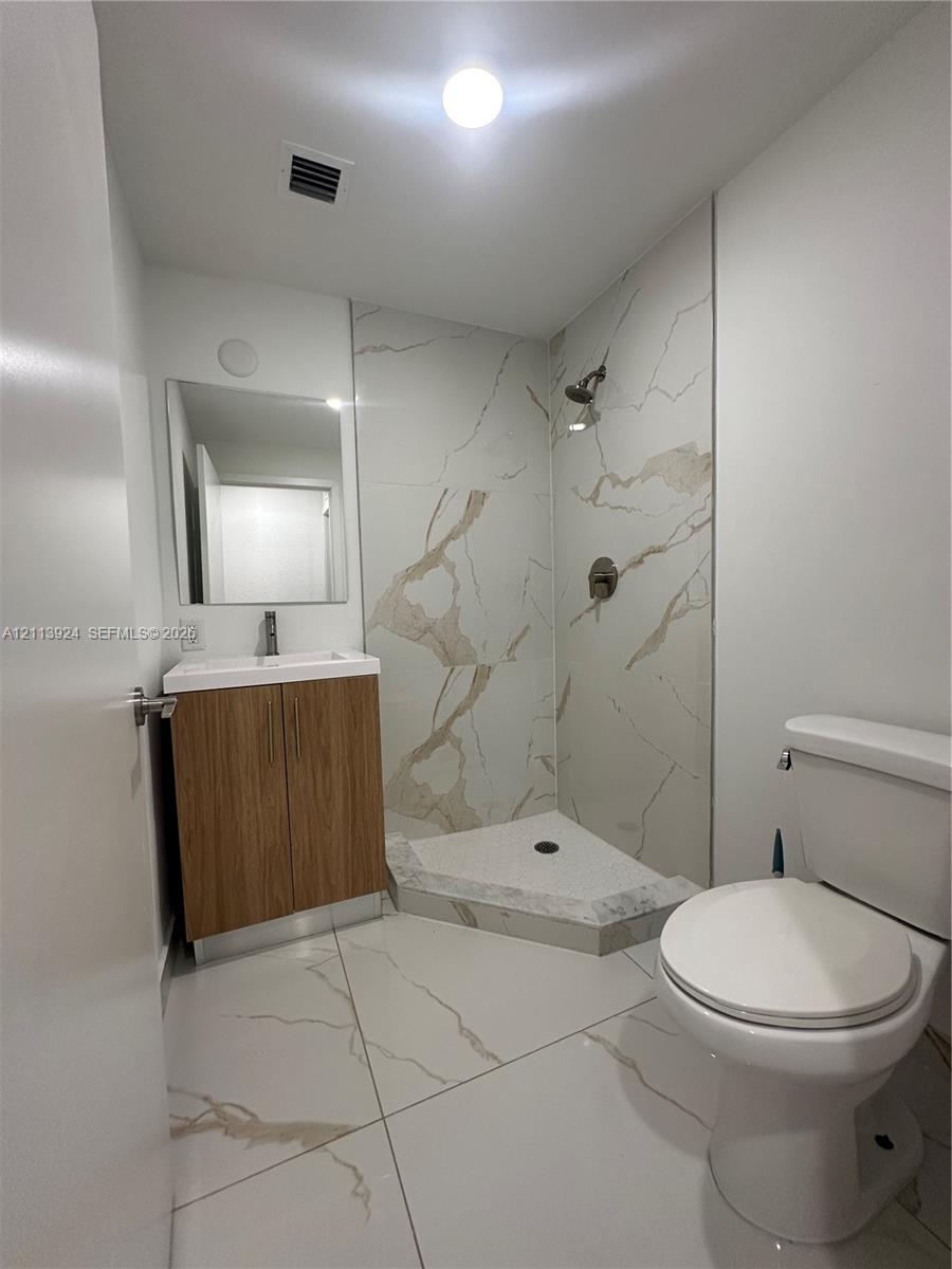 Bathroom featuring large-format veined tile flooring, a wood-finish vanity with a white basin, a corner shower enclosure with tiled surround, and a wall-mounted mirror