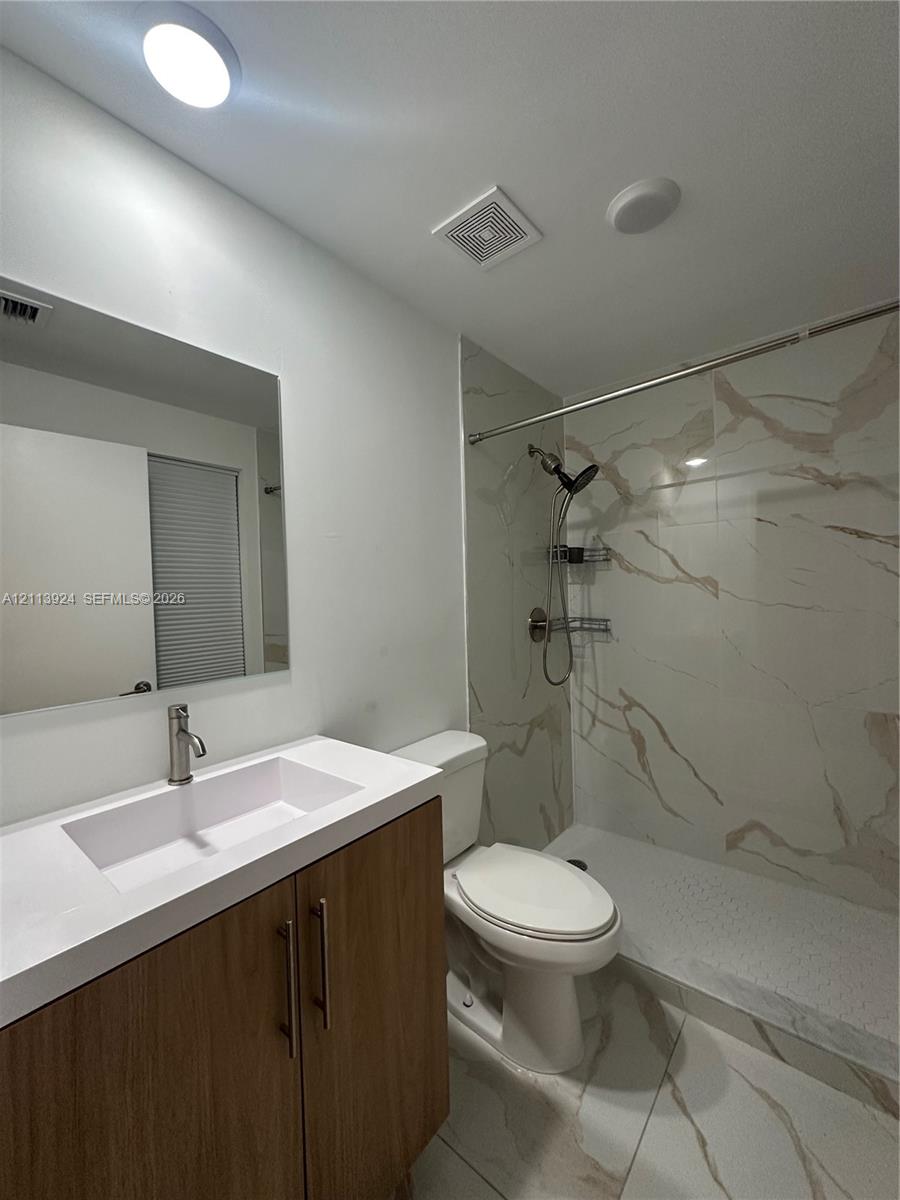 Bathroom featuring a wood-finish vanity with a white countertop and integrated sink, walk-in shower with floor-to-ceiling stone-patterned tile, and large-format tile flooring