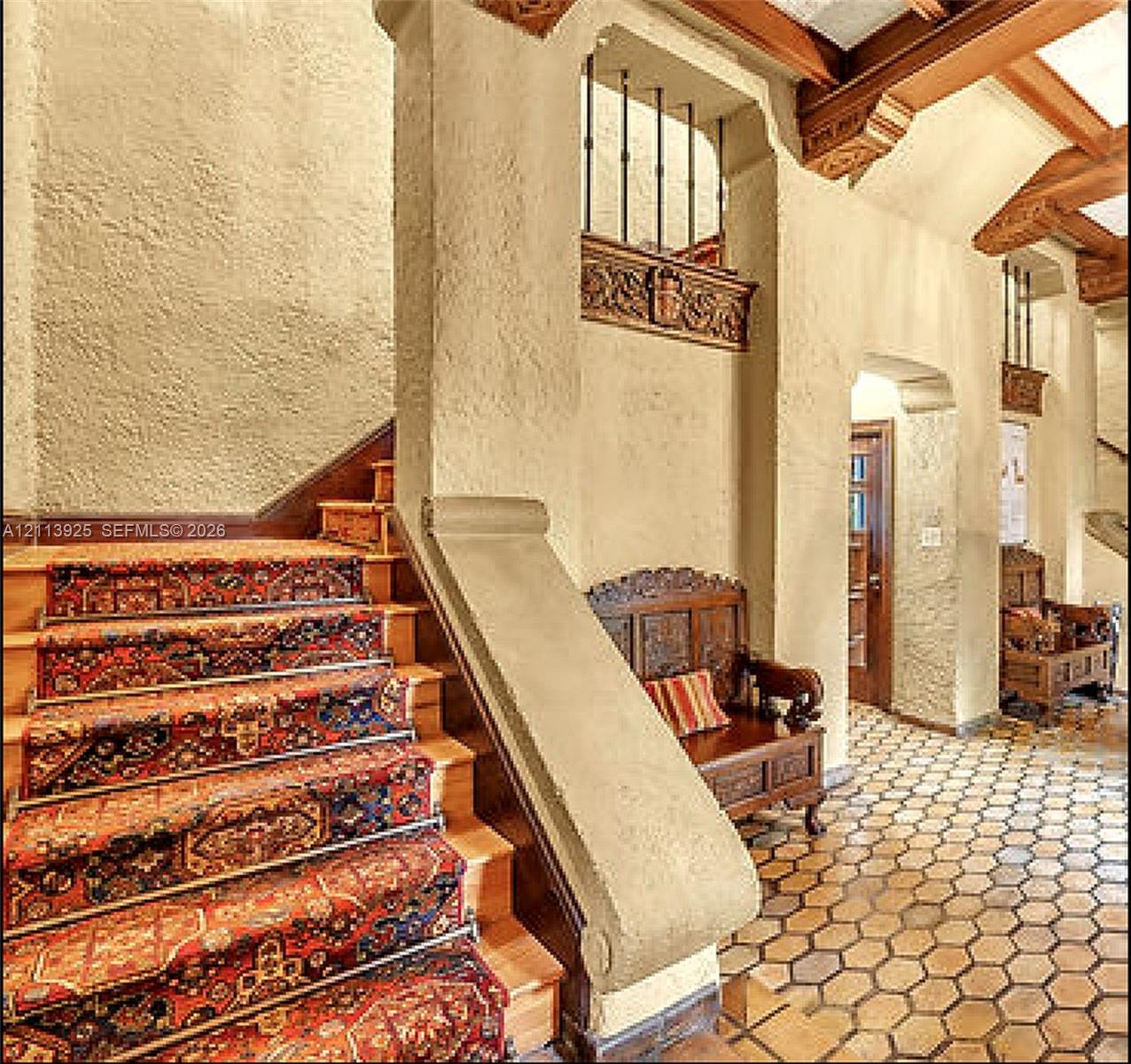 Staircase featuring wood treads and exposed ceiling beams, accented by detailed masonry archways and decorative carved wood trim, complemented by hexagonal terracotta tile flooring
