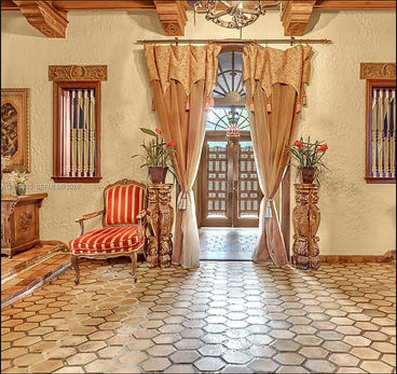 Entryway featuring hexagonal terracotta floor tiling, decorative wooden ceiling beams, dual casement windows with intricate carved wood surrounds, and a central arched entryway with decorative transom glass