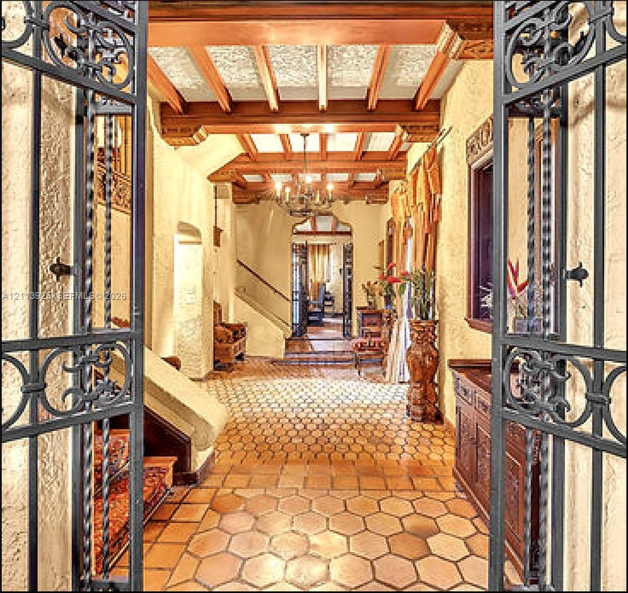 Foyer featuring intricate wrought-iron gate entry, hexagonal terracotta tile flooring, exposed beamed ceiling with decorative millwork, and a wrought-iron chandelier