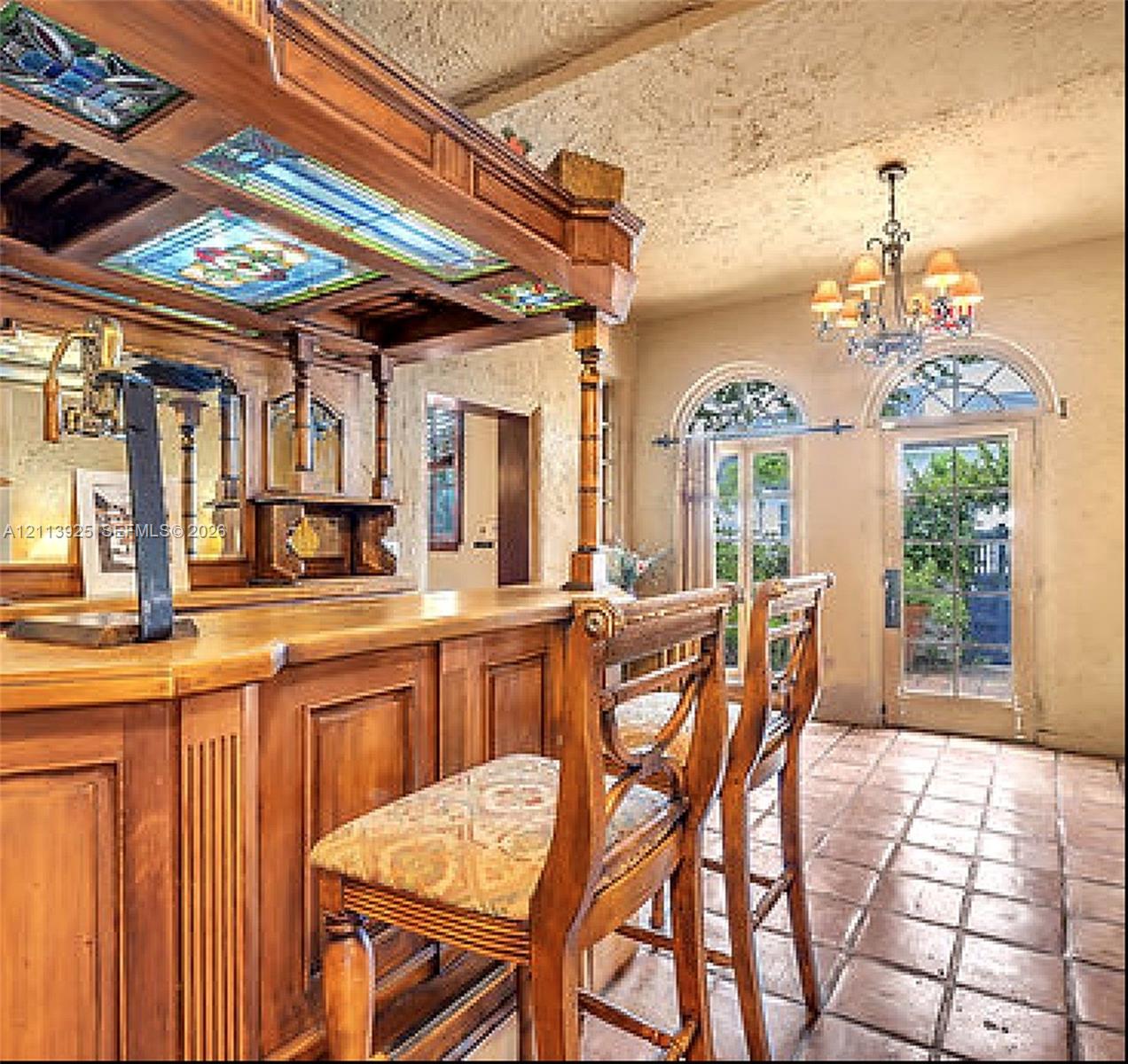 Custom wood-paneled bar featuring stained glass ceiling insets, ornate millwork, tiled flooring, arched glass-paneled doors, and a multi-tiered chandelier