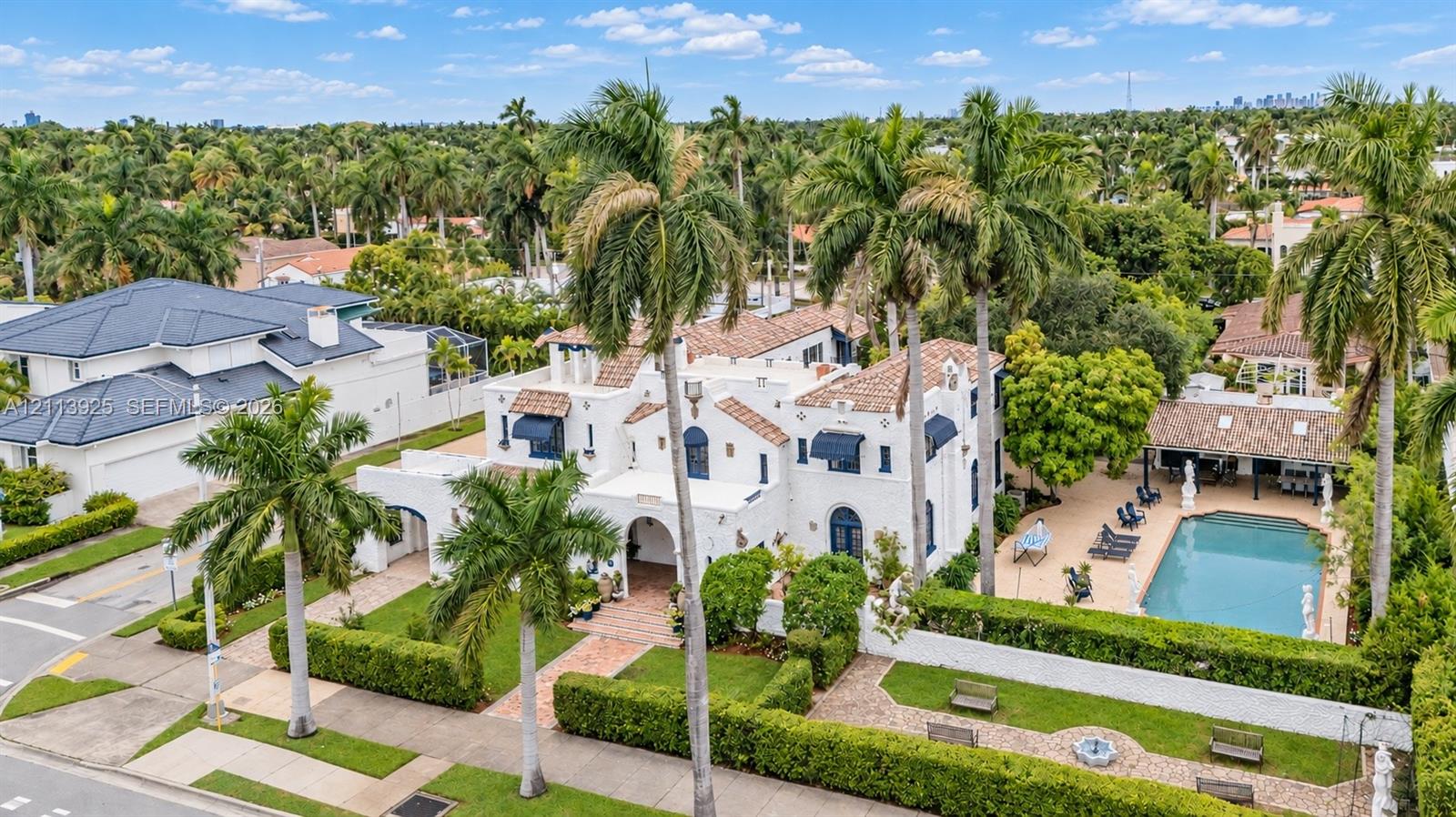 Spanish Colonial-style exterior with white stucco facade, tile roofing, arched entryway, private swimming pool, and landscaped grounds featuring mature palm trees