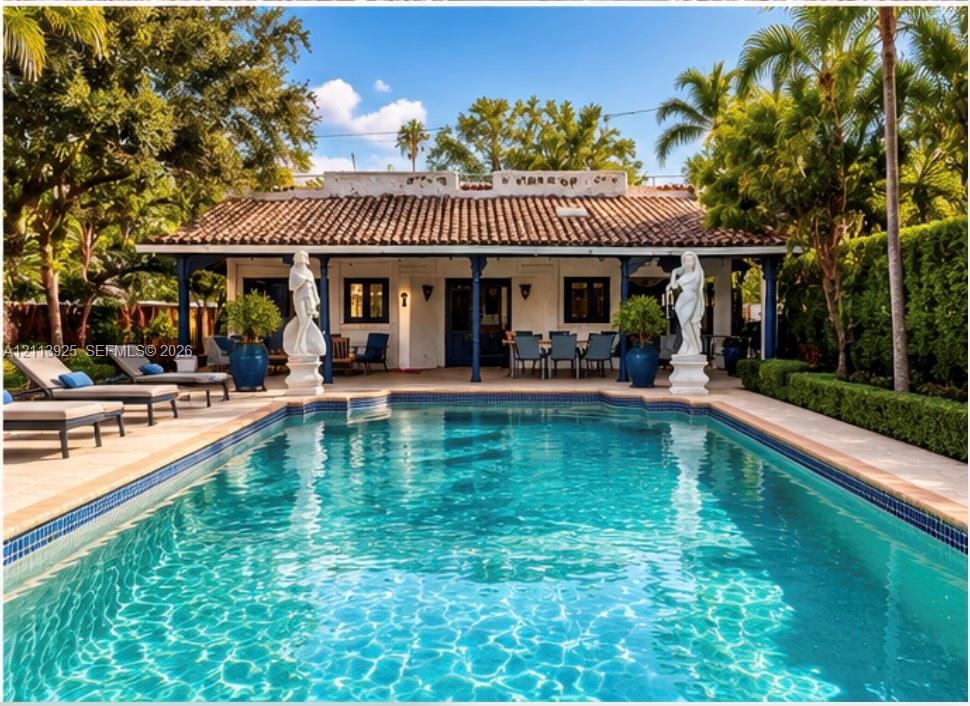 In-ground pool with decorative statues, expansive stone-paved patio, covered veranda featuring dark structural columns, and clay tile roof