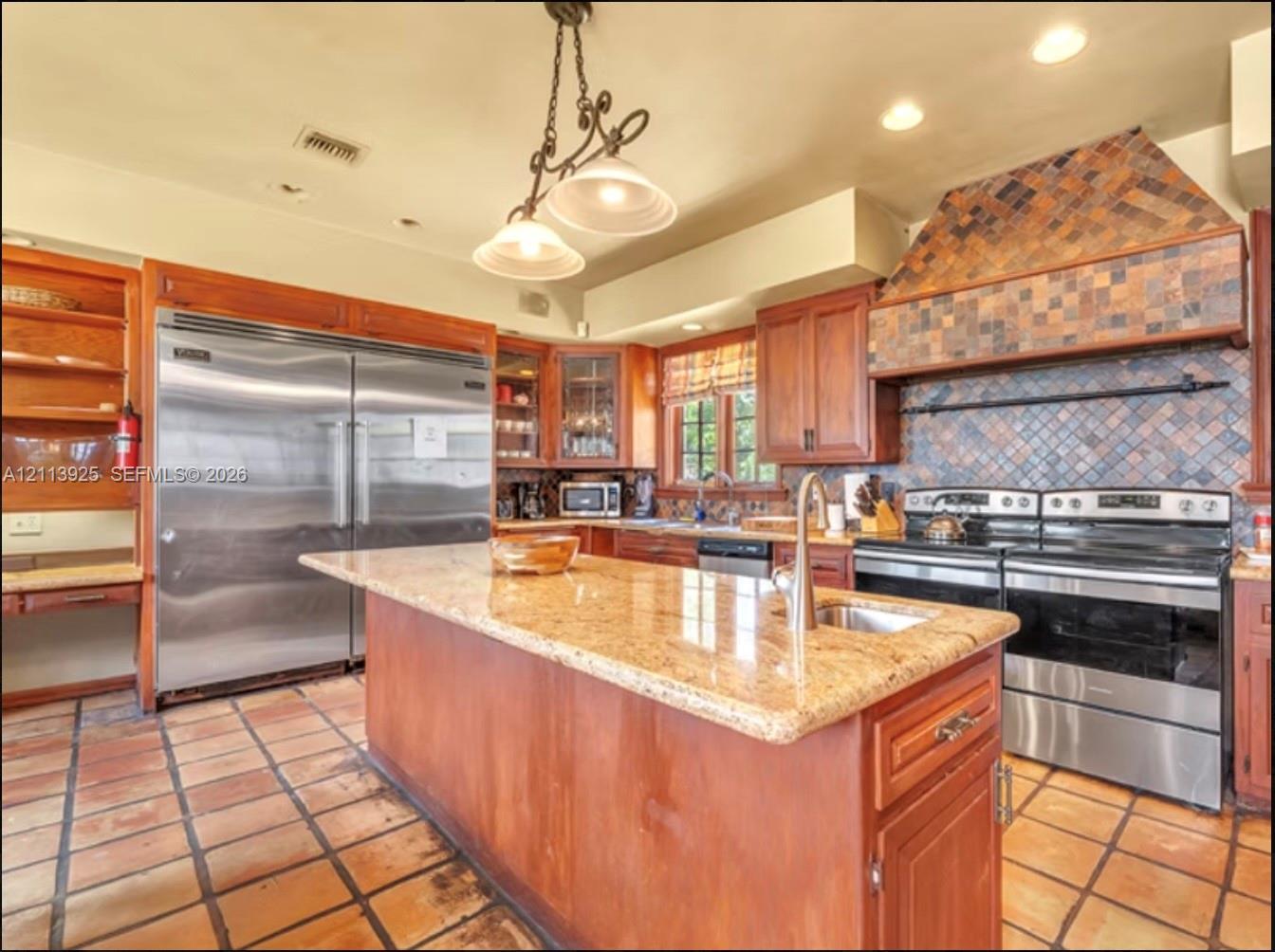 Kitchen featuring a large central island with stone-finish countertops, stainless steel appliances, custom wood cabinetry, a decorative tiled range hood, and terra cotta floor tiles