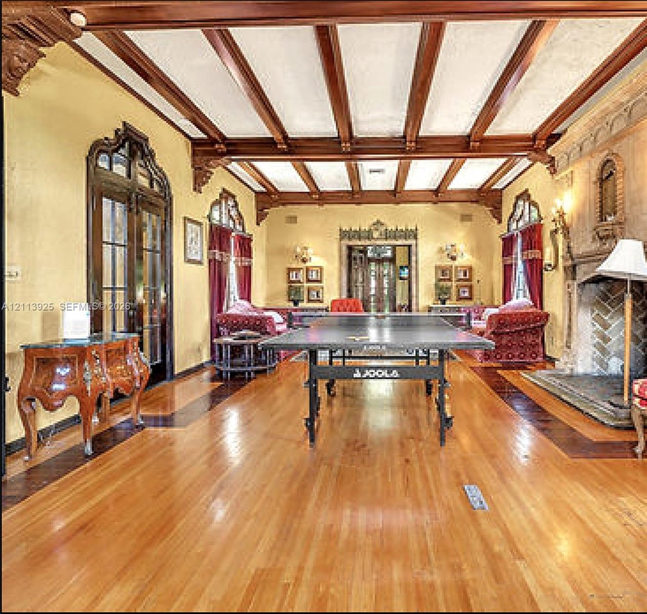 Expansive room featuring dark-stained wood ceiling beams, decorative corbels, wood-finish flooring with contrasting inlay borders, arched glass-paneled doors, and a floor-to-ceiling stone fireplace