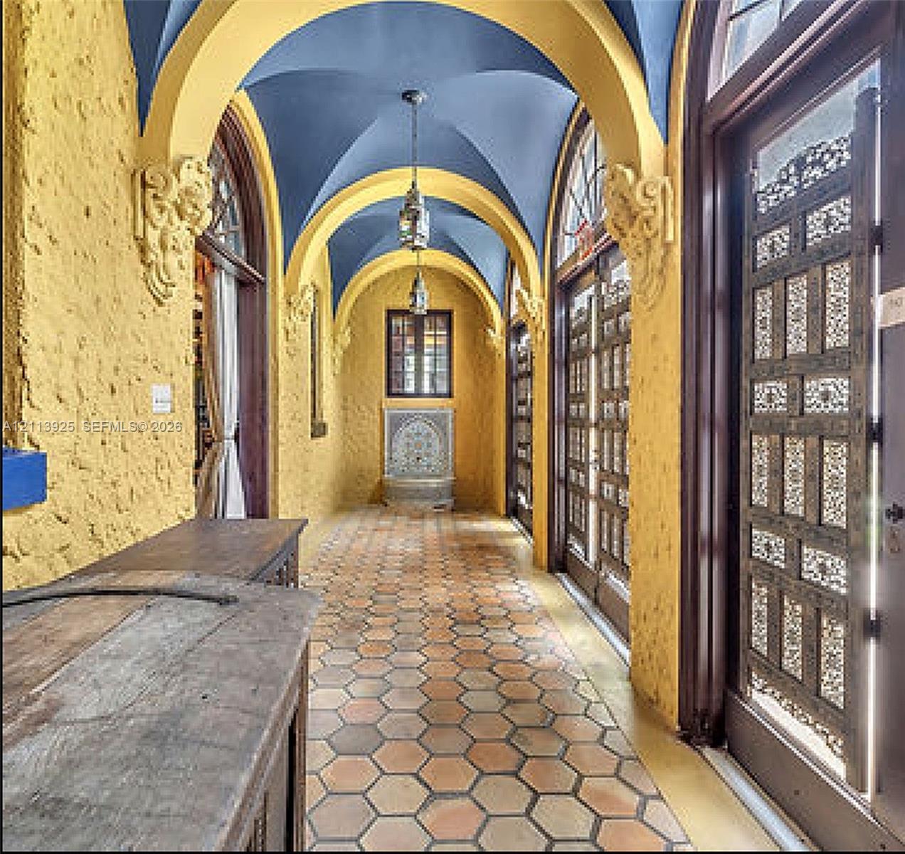 Vaulted corridor featuring a blue barrel-vaulted ceiling, decorative corbels, hexagonal terracotta tile flooring, ornate latticework doors, and an arched stone wall fountain