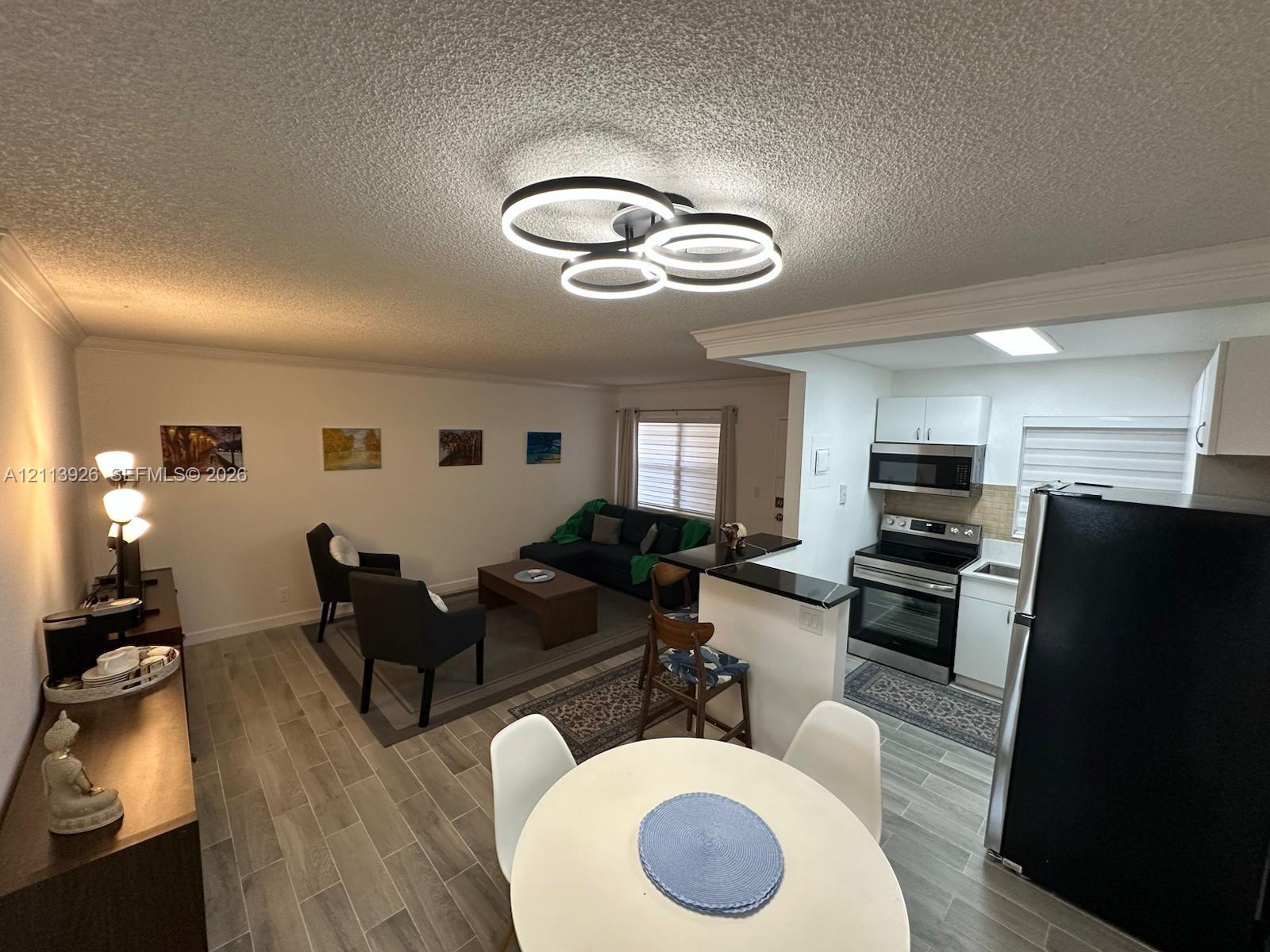 Contemporary multi-ring LED ceiling fixture, wood-finish flooring, and kitchen featuring dark stone-surfaced breakfast bar, stainless steel appliances, and white cabinetry