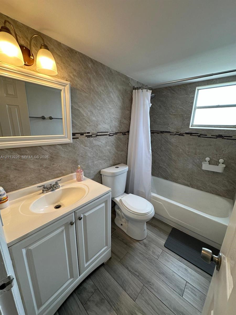Bathroom featuring wood-finish flooring, a white vanity with a molded basin, stone-finish wall tile, a decorative glass tile accent border, and a tub-shower combination